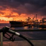 Oil tanker at an oil terminal at dusk with a fuel nozzle in the foreground, illustrating rising oil prices